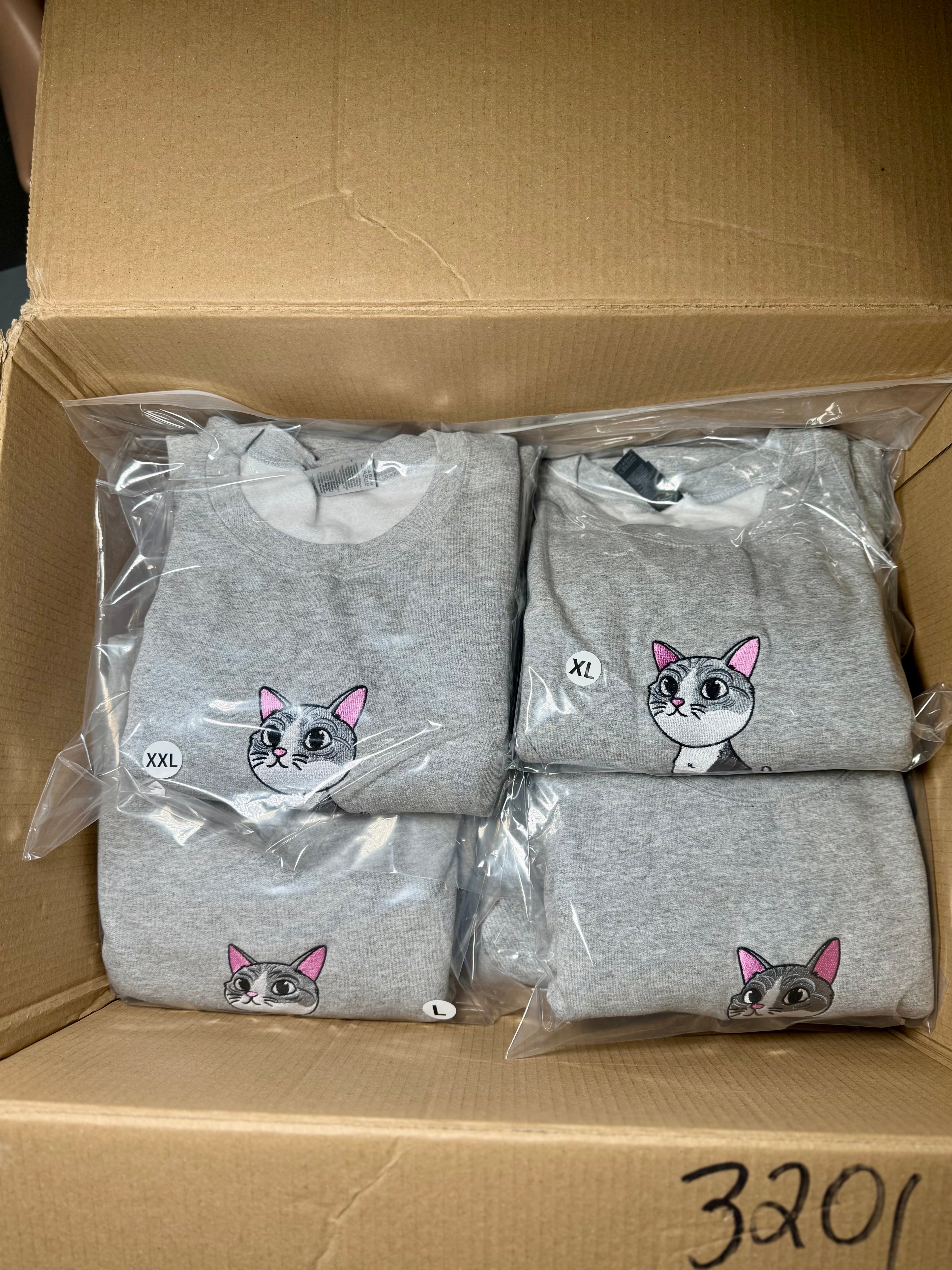 Three gray sweatshirts with cat designs in clear packaging inside a cardboard box.