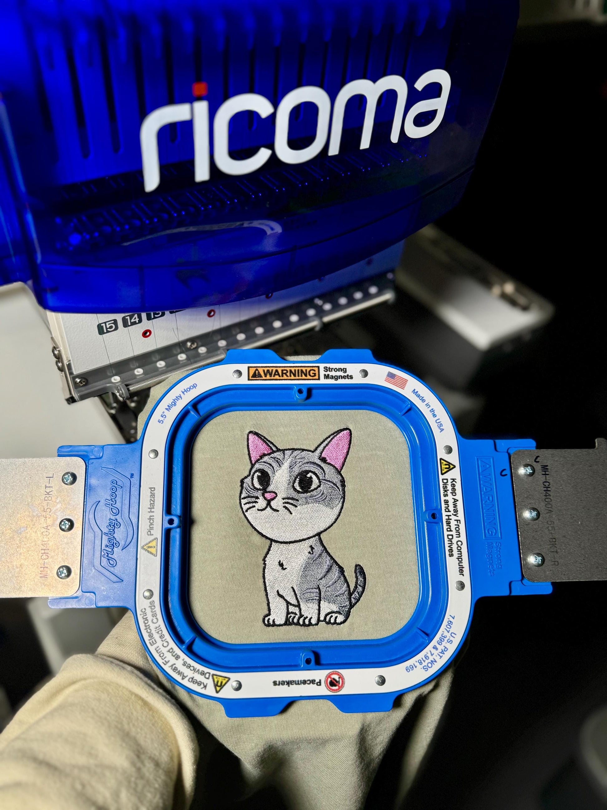 Embroidery machine with a cat design on a piece of fabric, brand 'ricoma' visible.